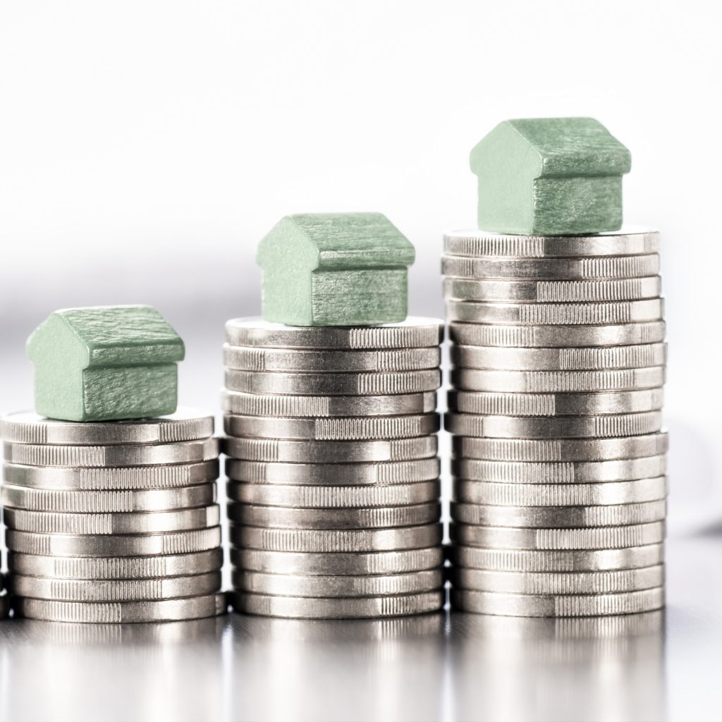 Kleine grüne Häuser stehen auf Stapeln aus Münzen mit Bauplänen im Hintergrund.